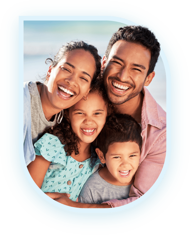 A happy family, consisting of a mother, father, daughter, and son, hugging and smiling together against a beach backdrop| Pleasant Dental