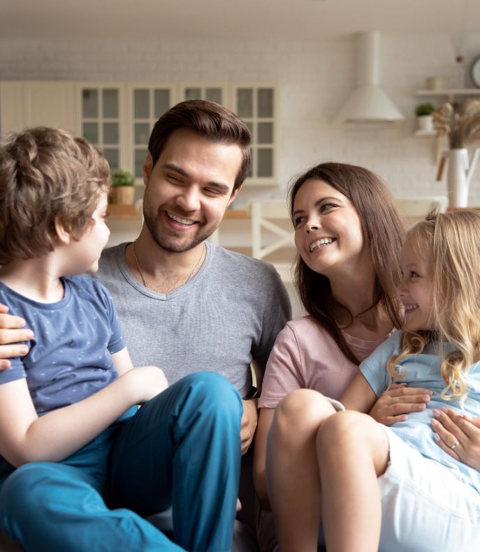 A smiling family sitting together at home, with a mother and father enjoying a moment with their young son and daughter in a brightly lit living room | Pleasant Dental